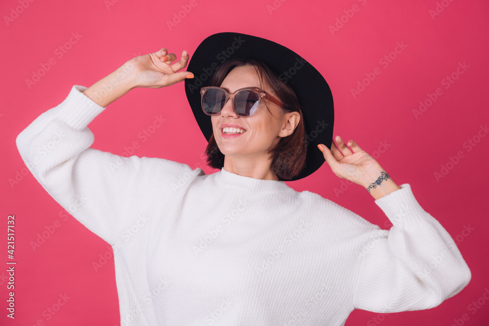 Wall mural stylish woman in white casual sweater, sunglasses and hat on red pink background copy space smile po