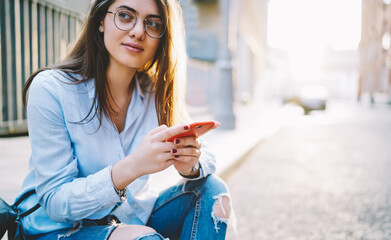Contemplative female in optical spectacles resting in city holding smartphone and thinking about online chatting, thoughtful hipster girl using mobile phone at urban setting pondering on network text