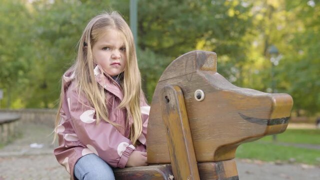 Cute little Caucasian girl frowns at the camera as she sits on a rocking horse at a playground in a park
