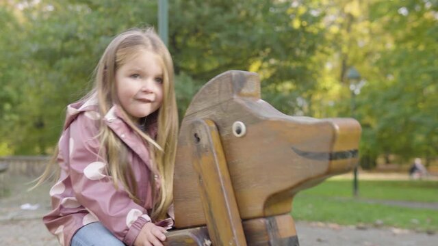Cute little Caucasian girl rocks on a rocking horse and smiles at the camera at a playground in a park
