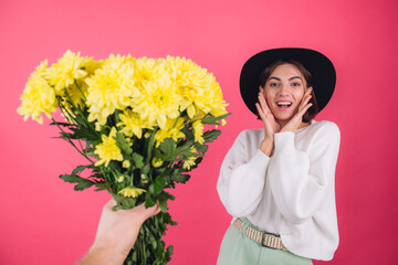 Stylish woman in hat and casual white sweater on pink red background gets large bouquet of yellow flowers, spring mood, isolated space amazed excited surprised