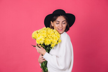 Stylish woman in hat and casual white sweater on pink red background hold bouquet of yellow flowers, spring mood, isolated space happy smile