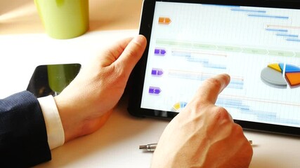 Business man scrolling tablet with the hand, showing a gantt for project management purposes. Smartphone and pen on the desk