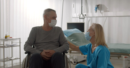 Female nurse wearing surgical mask talking to male patient sitting in wheelchair in hospital ward