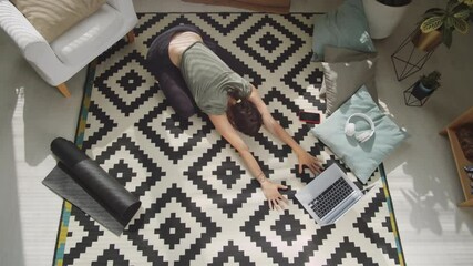 Top down shot of young slim woman in sportswear resting in extended child’s pose on floor at home while practicing yoga with online class on laptop