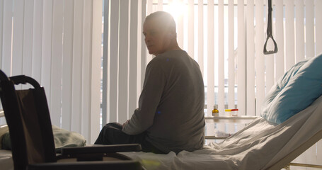 Senior disabled man sitting on hospital bed and looking at wheelchair