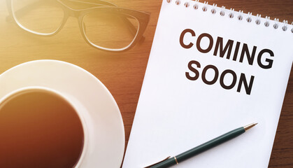 COMING SOON - text on paper with cup of coffee and glasses on wooden background in sinlight.