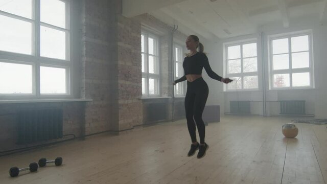 Slow-motion full shot of young fit caucasian woman in tight black sportswear jumping rope while training in smoky loft style gym studio alone