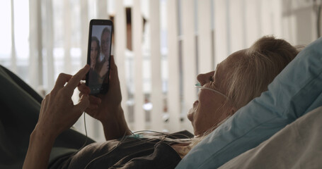 Senior female patient lying in hospital bed and having video call with family on smartphone