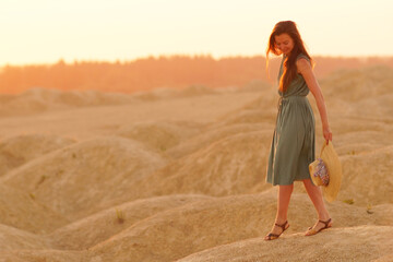 Young beautiful smiling woman with long hair in long blue dress with straw hat in hand on sand at sunrise in desert