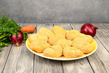 Raw cutlets in breadcrumbs lie on a white dish. Greens, carrots and a couple of bulbs are visible in the background