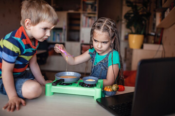 Sister and brother watching video on the screen of laptop and playing in master chef, preparing dinner with toy vegetables and real fresh eggs, online cooking during lockdown, at-home kid activity