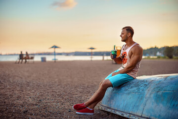 Fototapeta na wymiar Young handsome sporty man in bright clothes sits on the seashore, drink a refreshing cocktail and enjoys his vacation.