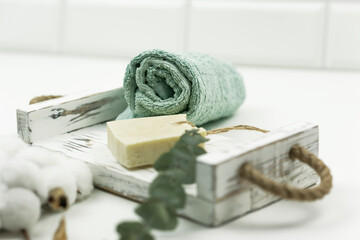 A white scrub hand made soap, a cotton towel, a branch of cotton and green eucalyptus lie on a white wooden tray