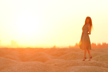Beautiful woman with long hair in blue dress with straw hat in hand standing on sand in desert at sunrise, backlight