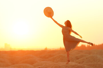 Elegant woman with long hair in dress standing on one leg in sandy desert, straw hat raised in hand, sunrise, backlight