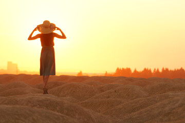 Elegant woman in long dress with straw hat standing on sand in desert, back view. Sunrise, backlight photo