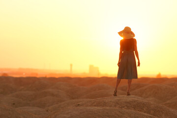 Elegant woman in long dress with straw hat standing on sand in desert, back view. Sunrise, backlight photo