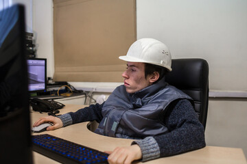 An engineer in a white helmet sits in front of the monitors. A young systems engineer works in his office. Portrait of a male designer.
