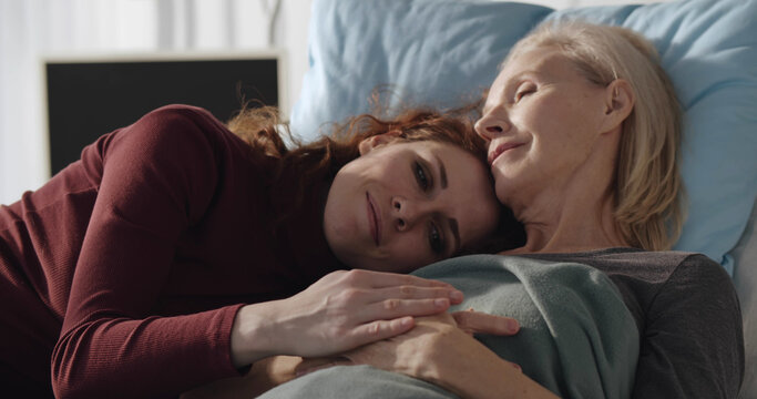Young woman lying in hospital bed with sick aged mother and hugging during visit in clinic