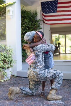 African american soldier father hugging smiling daughter holding flag in front of house