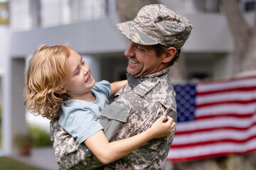 Smiling caucasian soldier father carrying son in garden with american flag hanging outside house