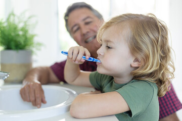 Smiling caucasian grandfather in bathroom kneeling beside grandson brushing teeth