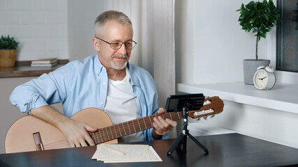 An elderly male blogger teaches guitar playing in an online lesson at home.