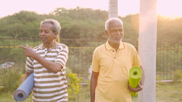 Fron View Follow Shot Of, Two Senior Men By Holding Yoga Mat Coming To Park With Backlit Sunlight- Older Joyful Friends Walking And Talking In Garden During Morning After Workout