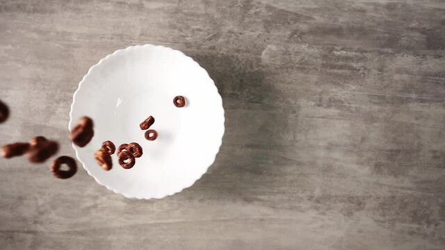 Chocolate cereal rings pouring into white plate standing on wooden table in slow motion. Cooking delicious morning meal. Traditional breakfast made of ready to eat food. Cornflakes falling from above.