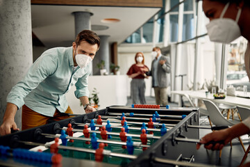 Businessman with face mask playing table football with colleague while taking break from work in...