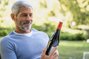 Smiling mature man looking at wine bottle while sitting on chair