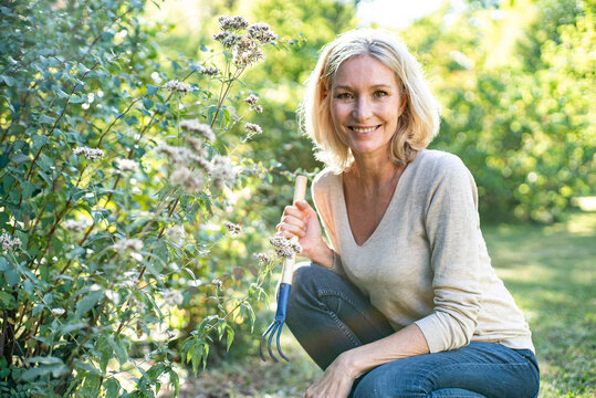 Portrait of smiling mature woman with garden fork crouching in backyard