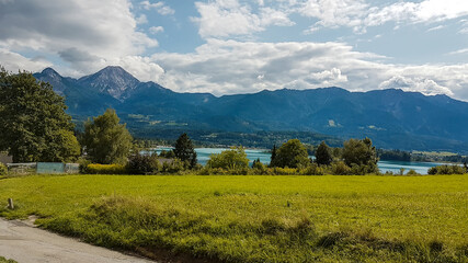 A distant view on the Faaker lake in Austrian Alps. The lake is surrounded by small buildings and tall trees. In the back there are high mountains. Lush green meadow in front. Spring in the mountains.