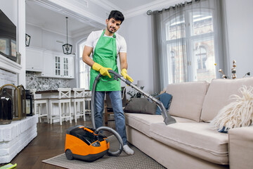 Fototapeta na wymiar Indian guy in uniform cleaning couch with modern vacuum