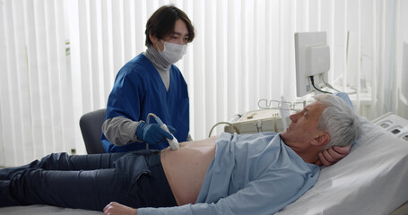 Doctor wearing safety mask with ultrasound scan examining abdomen of senior male patient in clinic