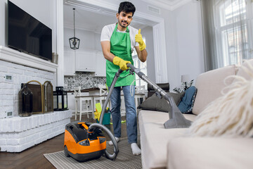 Indian young man cleaner in uniform cleaning sofa with modern vacuum