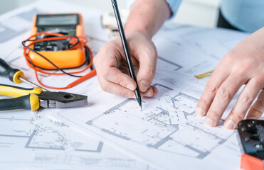 Man,repairer making electricity project in house.Repairs planning.Drawings,diagrams,plan for electrification of apartment,building.Devices,accessories,voltmeter,wires,screwdriver,pliers,tape measure - obrazy, fototapety, plakaty