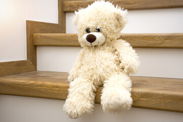 White plush teddy bear sitting on stairs at home and waiting. Lonliness, stress, depression of being alone at home concept.