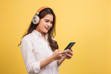 Smiling young woman using phone for listening to music on yellow background.