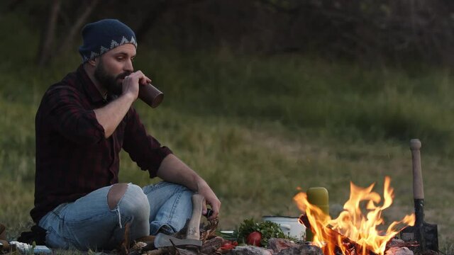 Thoughtful man tourist sits by campfire and looks into the blazing fire, drinks from bottle. Guy relax on outdoor tourism vacation, resting in his camp in wild plain area near woods