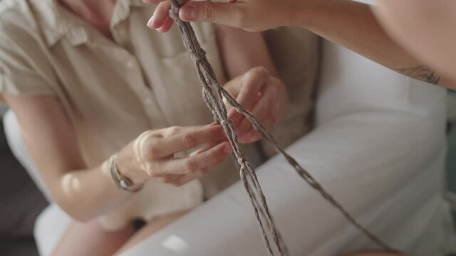 Dutch angle shot of young brunette woman knotting macrame plant hanger with help of female friend and speaking with her while spending day together at home