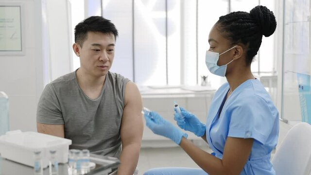 Young man communicating with doctor in protective mask and safety gloves while she filling syringe with vaccine in hospital while waiting for vaccination. Concept of prevention.