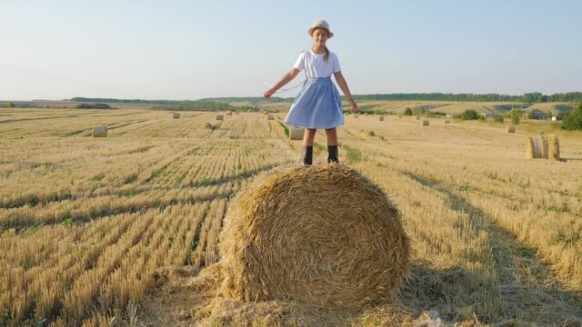 Happy childhood. A happy young girl in a dress and rubber boots is sitting on a haystack in a mown field. Beautiful little girl sitting on a haystack. Fun dream kid concept.