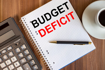 BUDGET DEFICIT written in a white notepad near a calculator and a cup of coffee on a dark wooden background