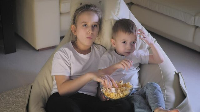 Cute little kids while watching TV. Two children watching TV, eating popcorn sitting on floor at home. Cute child family of two adorable siblings eating popcorn and watching cartoons in living room.