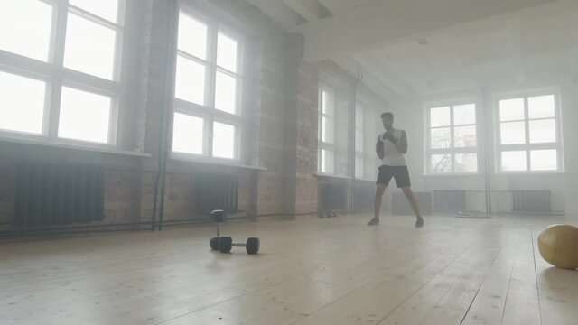 Slow-motion full shot of afro-american fit man training alone at loft style studio filled with chalk powder smoke practicing boxing punches with gloves