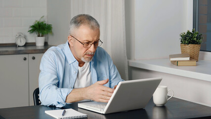 An elderly man with a beard feels furious when he looks at the laptop screen.