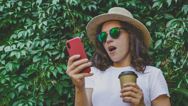 Excited young woman surprised by good unbelievable news, looking at smartphone screen feeling amazed, standing outdoors in green park, holding coffee cup to go. Unexpected win, shopping sale offer