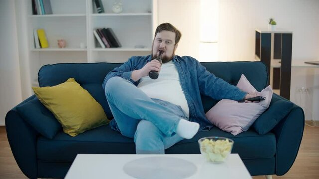 Lazy plump bachelor talking to tv, drinking beer on sofa, unhealthy lifestyle